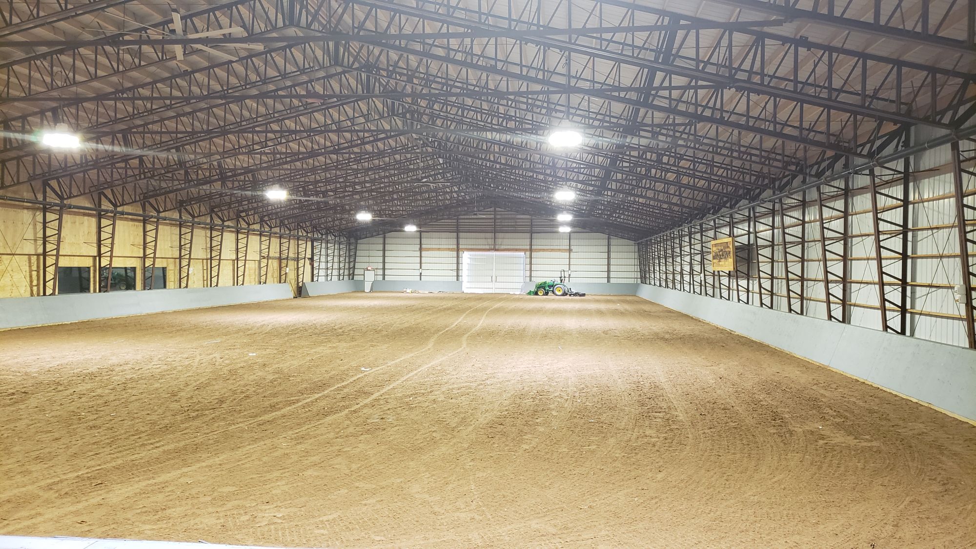 A spacious indoor horse riding arena with a dirt floor and a metal-beamed ceiling. Bright lights illuminate the space, and a green tractor is visible in the background. White walls line the arena sides, providing an ideal setting for equestrian activities.