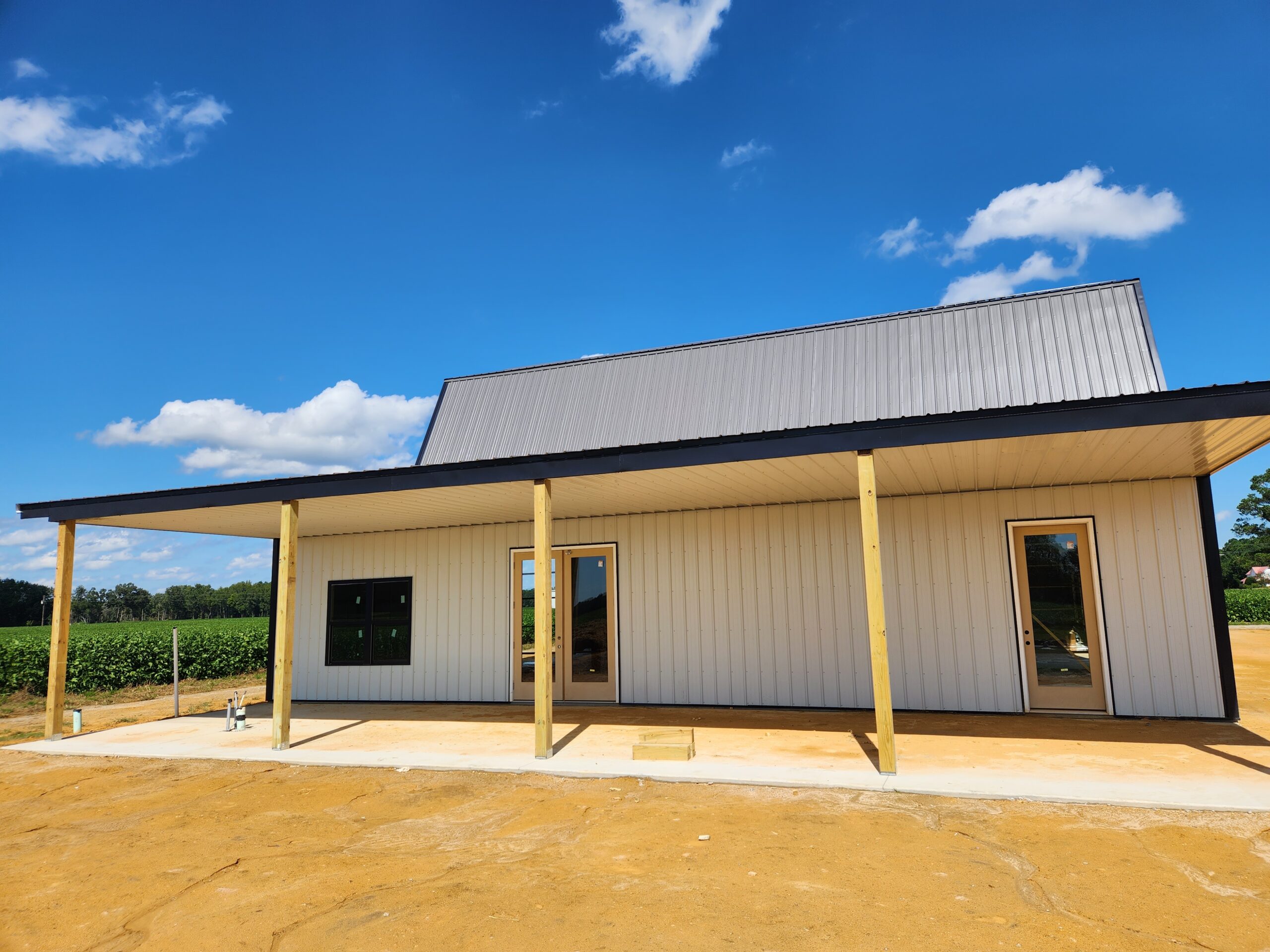 A modern barn-style house with a gambrel metal roof and wooden pillars, surrounded by a dirt lot. The house features large windows, a small covered porch, and an attached garage. It's a sunny day with a clear blue sky and a few white clouds.