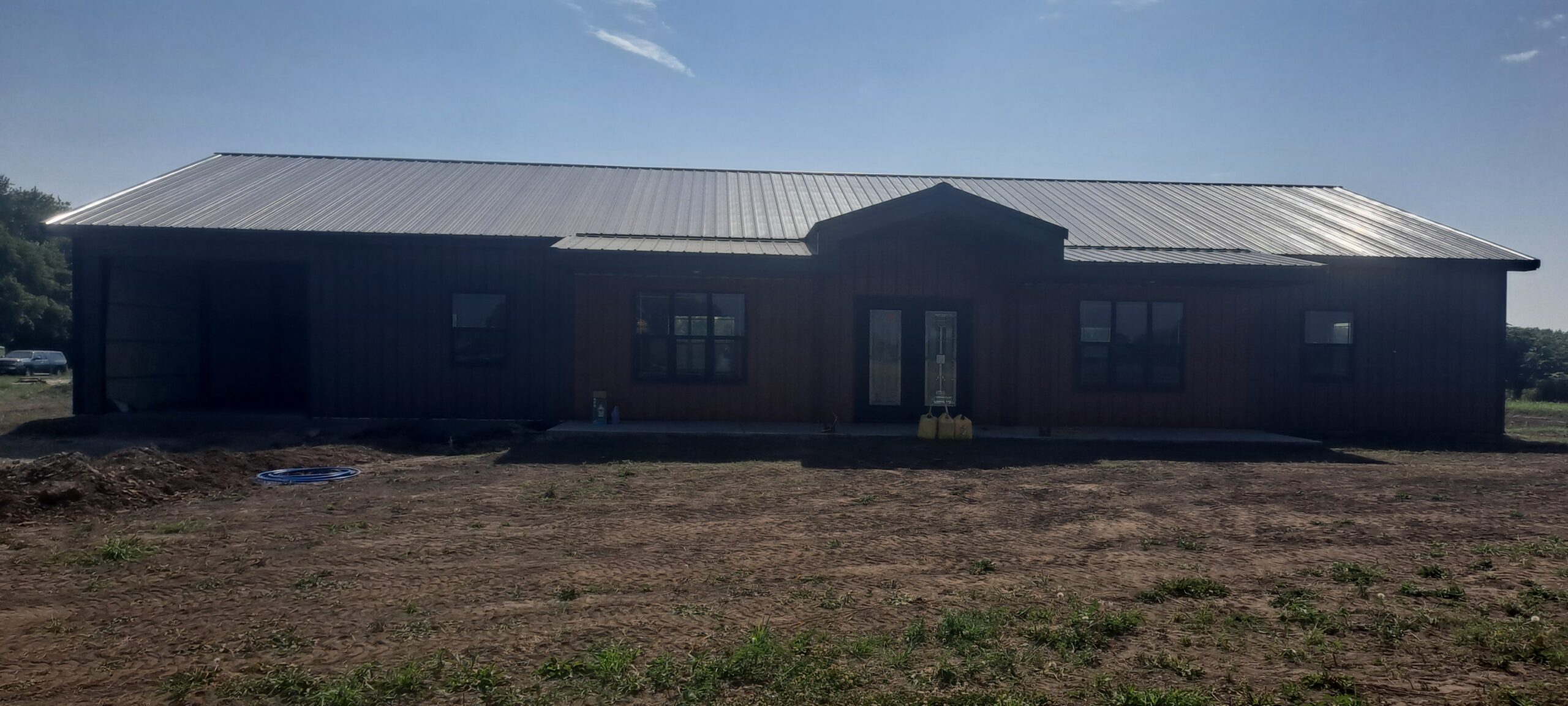 A large, single-story building with a metal roof and dark exterior sits on Reece Ranch's barren plot under a clear blue sky. This barndo features windows and double doors at the front, offering both functionality and charm in its rustic setting.