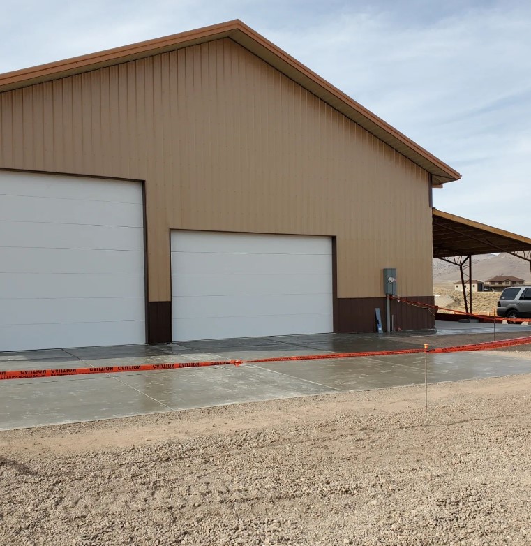 A large beige metal barndominium with two white garage doors stands in a desert setting. Caution tape stretches across the concrete driveway. To the right, a covered area and a sienna-colored SUV are visible under the clear, sunny sky.