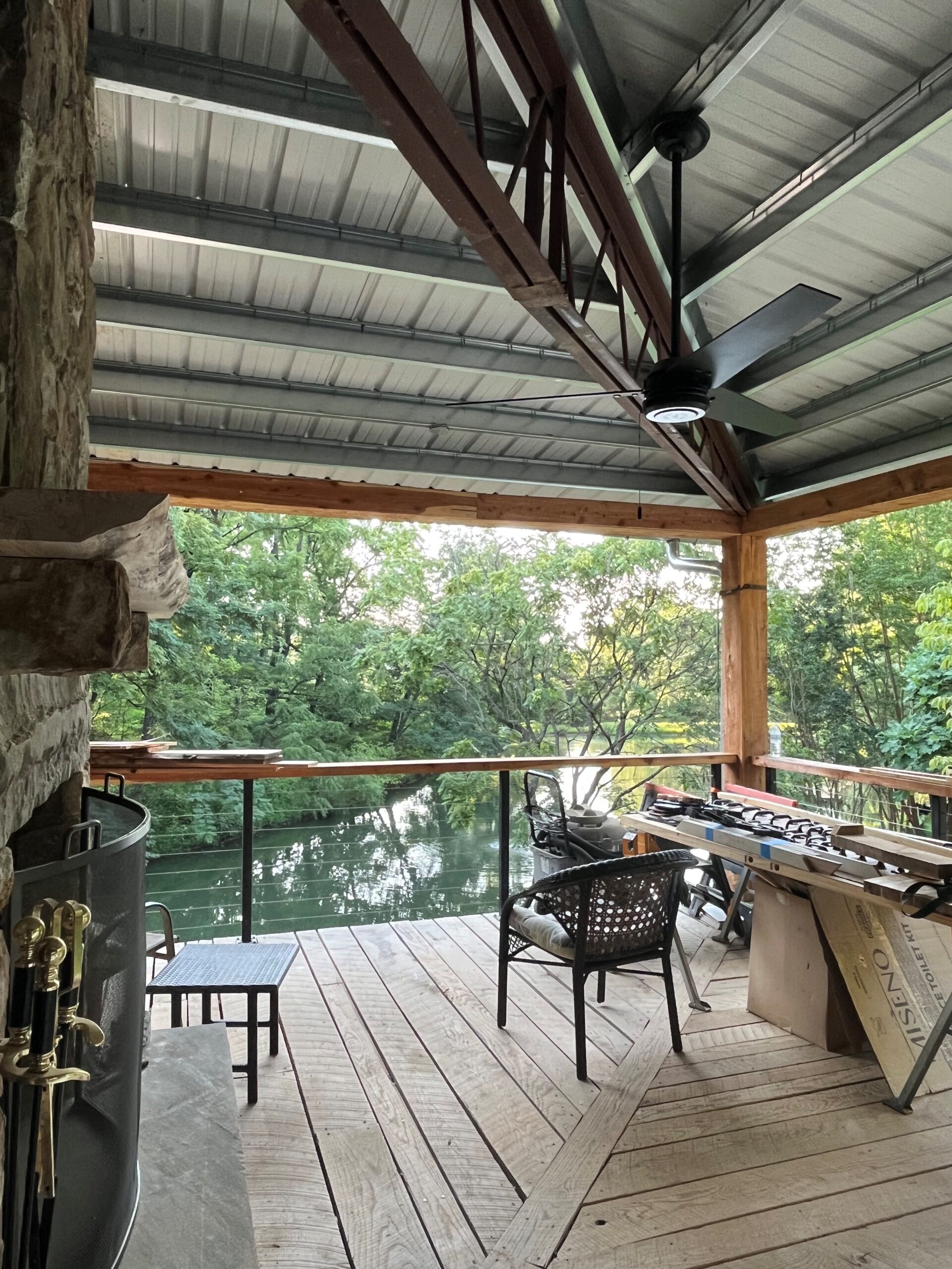 An asymmetrical wooden deck with a metal roof overlooks a serene river, embodying the charm of a rustic lakehouse. It features wicker chairs, a ceiling fan, and a fireplace, with tools scattered on the table beneath lush green trees. The atmosphere is peaceful and inviting.