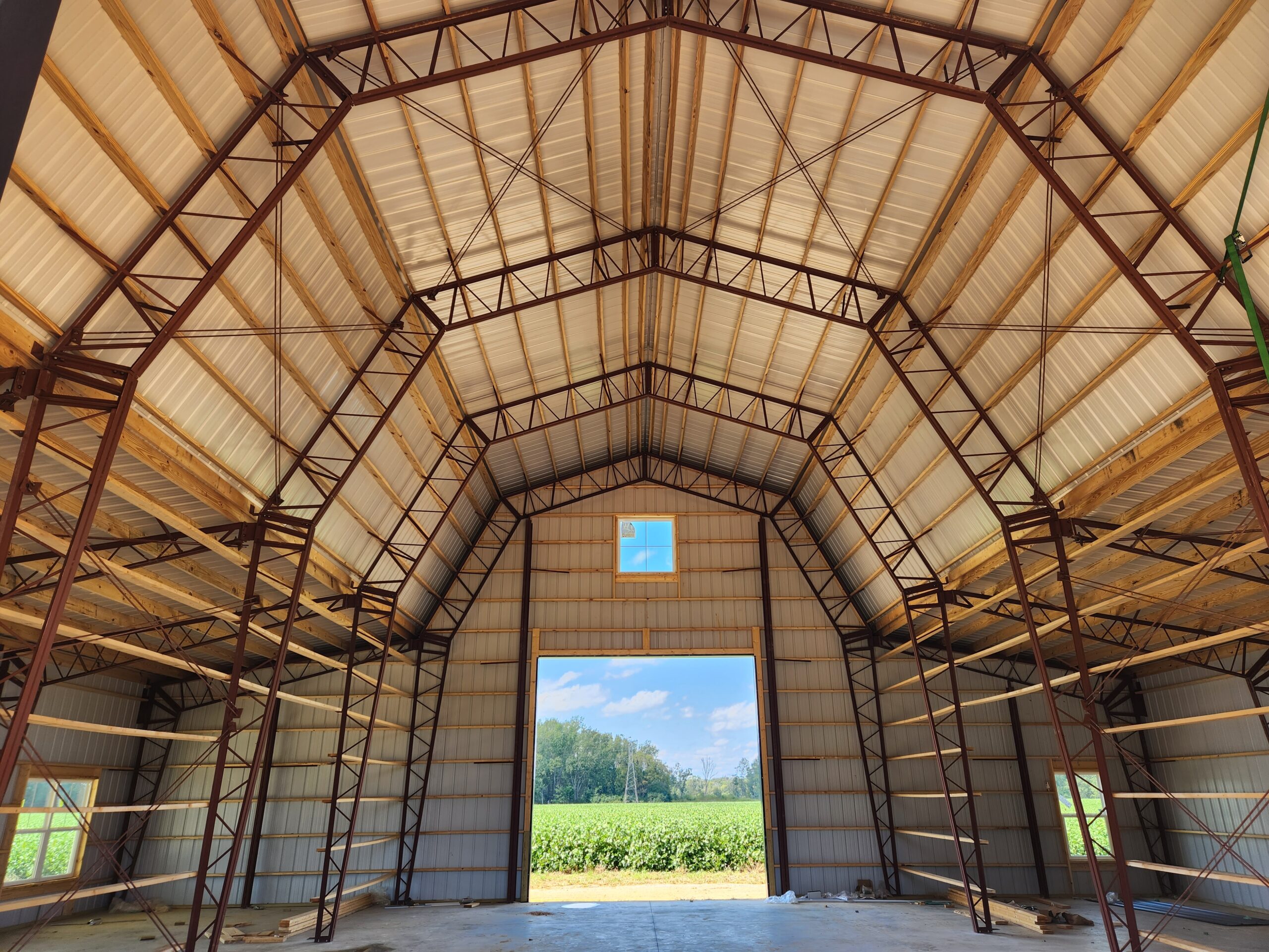 Interior view of a large, empty barn with a high metal gambrel roof and sturdy beams. Sunlight shines through the open barn door, revealing a view of a lush green field and trees in the distance.