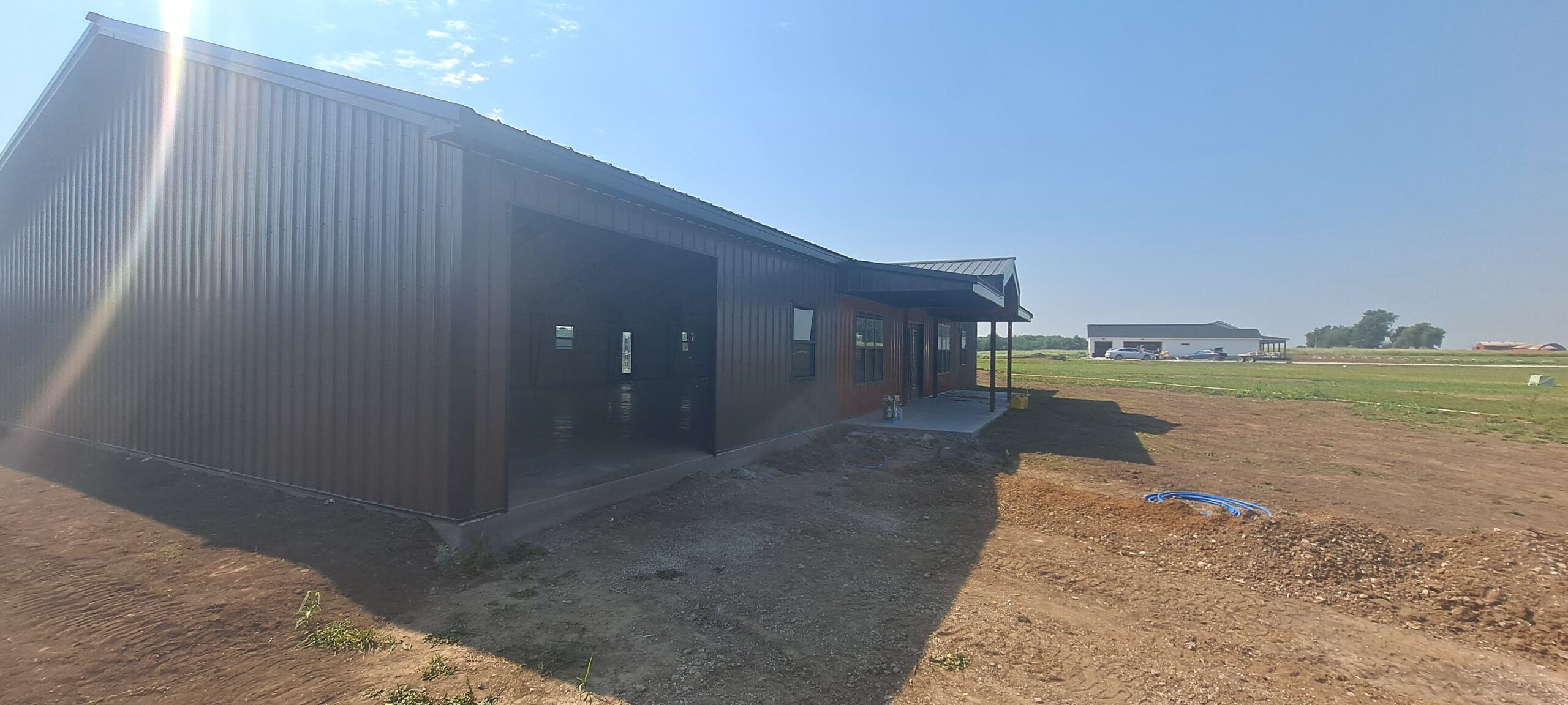 A large, metal barn-like structure, reminiscent of a Reece Ranch Barndo, stands with an open entrance on a grassy field under a clear blue sky. Nearby, another building is visible. The foreground showcases dirt and patches of grass.