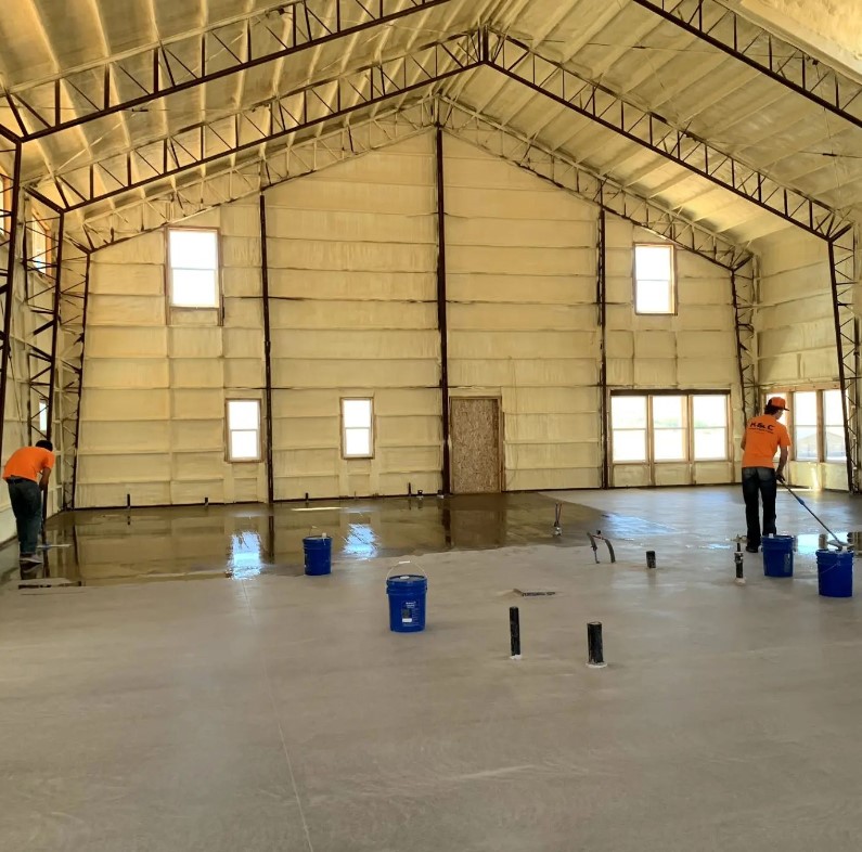 Workers in sienna orange shirts are smoothing a concrete floor in a barndominium-style warehouse with high ceilings and exposed steel beams. Several blue buckets are scattered on the floor, and large windows line the walls.