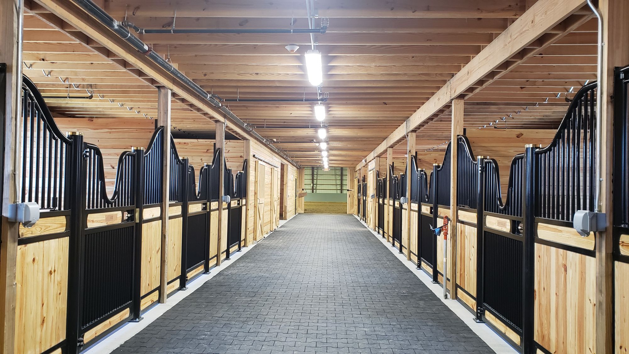 A clean, well-lit horse stable aisle with wooden stalls on both sides evokes the ambiance of a riding arena. The ceiling and walls are crafted from wood, complemented by metal gates at each stall entrance. Rubber mats pave the floor, leading to a door at the end of this inviting space.