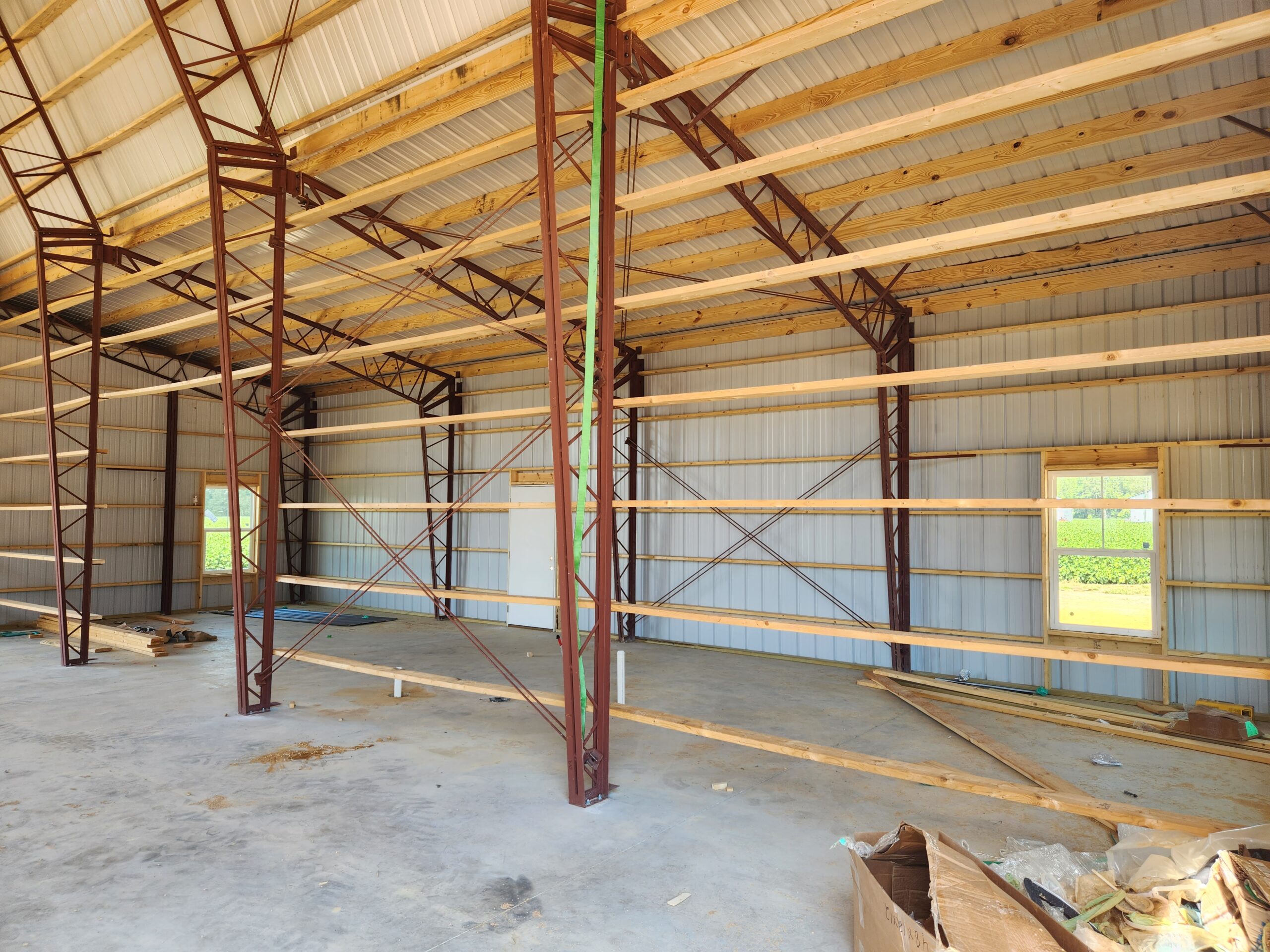 Inside the expansive warehouse, reminiscent of a vast garage with its high ceilings and metal framework, sunlight streams through the side windows. Wooden beams support the structure, while construction materials are scattered across the floor in anticipation of transformation.