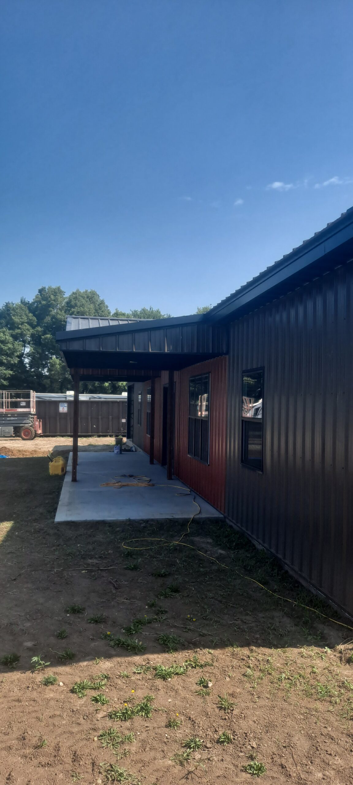 The image depicts a barndo-style modern building with a metal exterior and sloped roof, reminiscent of Reece Ranch. An overhang shelters a small concrete patio, while the grassy surroundings and scattered trees bask under a clear blue sky.