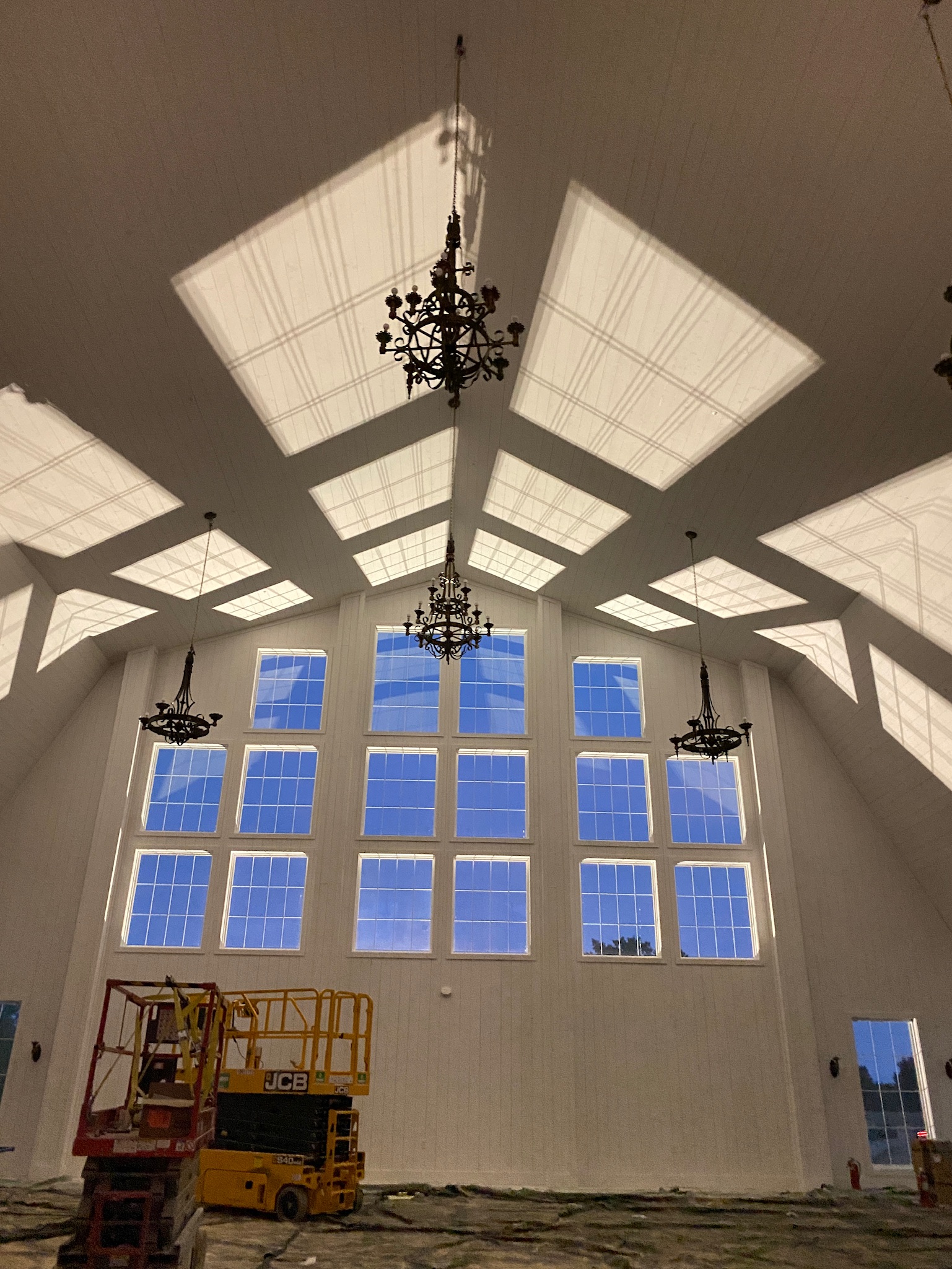 The interior of a grand wedding venue features high ceilings adorned with ornate chandeliers. Sunlight streams through tall windows, casting patterns on the walls. Scaffolding and other construction equipment suggest that work is still in progress to perfect this majestic gambrel-roofed setting.