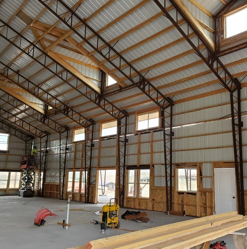 The interior of the large barndominium under construction showcases high ceilings and a robust metal framework, with partially installed windows filtering warm sienna light. Construction materials and tools are scattered across the concrete floor, hinting at the industrious activity underway.