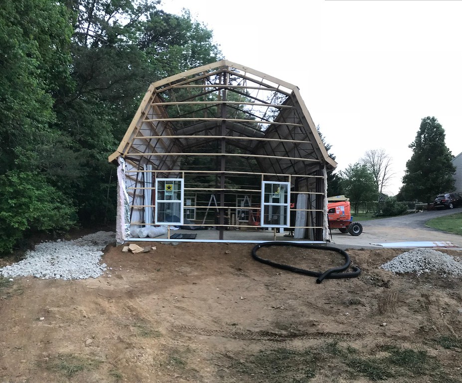 A partially constructed barn structure with a gambrel roof stands on a dirt and gravel foundation. Its wooden frame is visible, with metal panels on the roof. Trees surround the area, and a red construction lift is parked nearby. The sky is overcast.