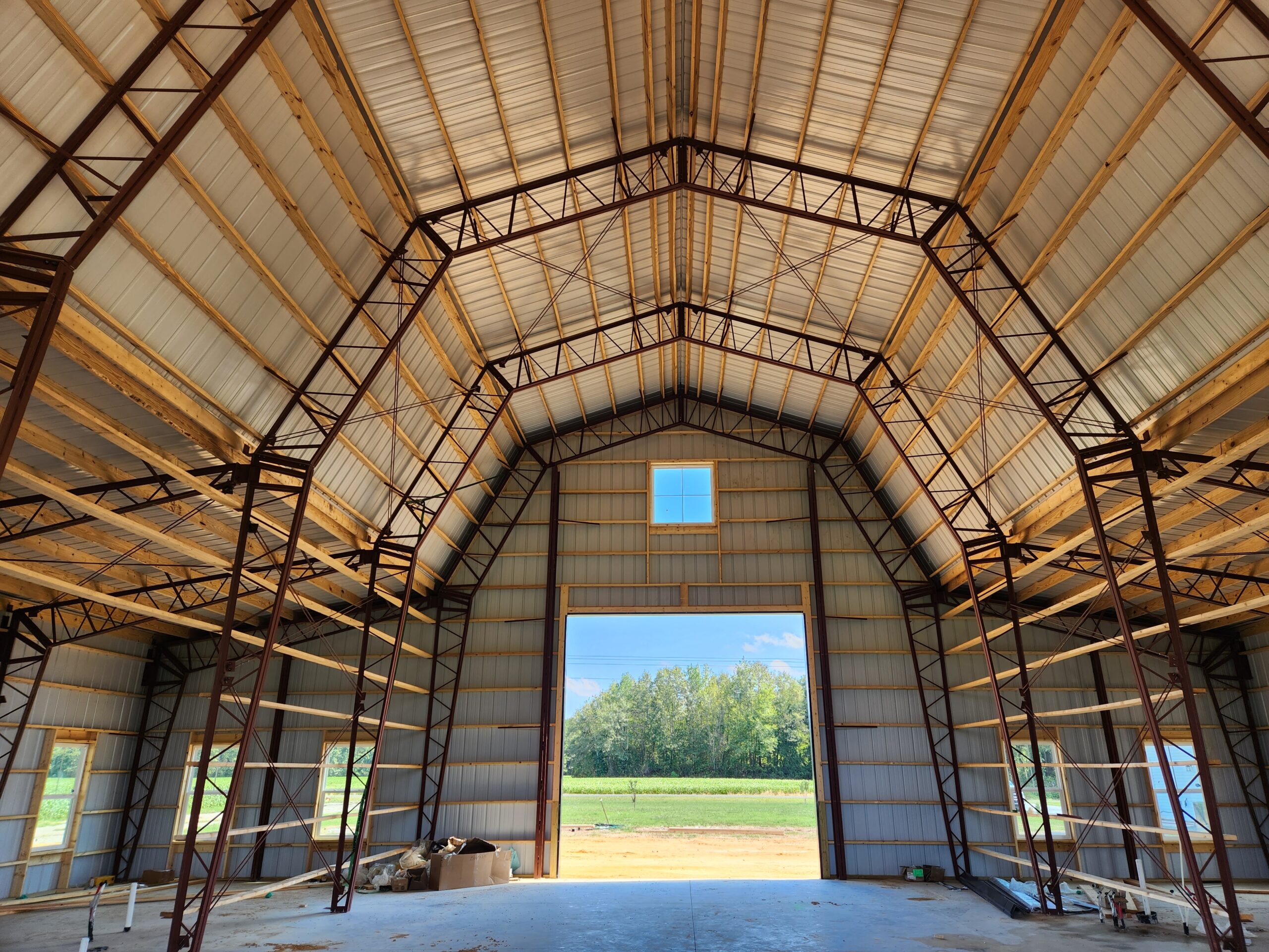Interior view of a large, empty metal barn with a high ceiling and visible steel beams, reminiscent of a gambrel style. Sunlight streams through the wide-open door, revealing a grassy field and trees outside. Construction materials are scattered across the floor.