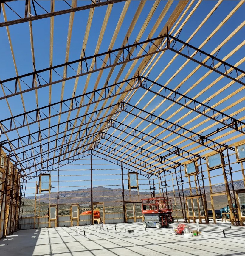 Interior view of a large, unfinished steel-framed barndominium with wooden beams under a clear Sienna sky. Construction equipment is visible on the concrete floor, and mountains can be seen in the background.