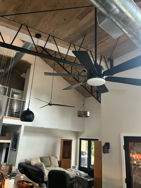 The lakehouse living room boasts an asymmetrical design with a high wooden ceiling and exposed metal beams. A large black ceiling fan and modern pendant lights complement the cozy sofa below the loft area. Natural light streams in through glass doors, creating a serene ambiance.
