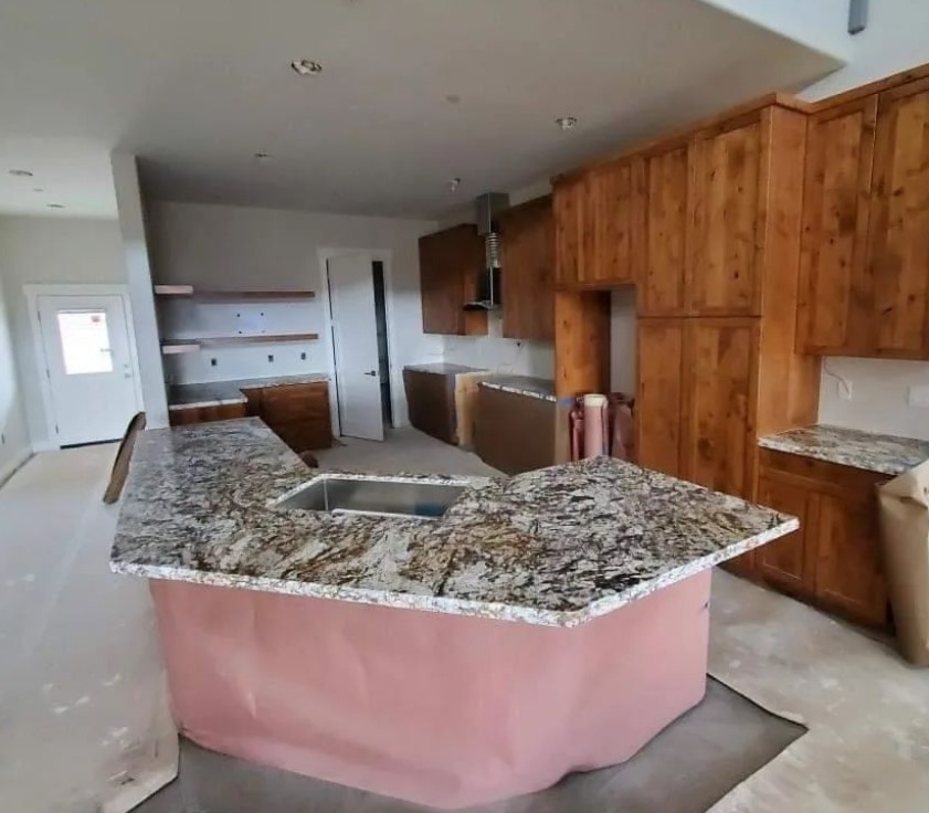 A modern kitchen under construction within a Sienna Barndominium features a sleek granite L-shaped countertop. Wooden cabinets are installed along the walls, and the floor is partially covered for protection. Light pours in through a door in the background.