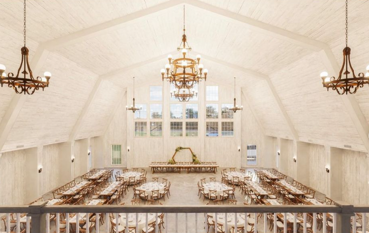 This charming wedding venue features a spacious banquet hall with high gambrel ceilings, large windows, and wooden chandeliers. The setting boasts numerous wooden tables and chairs in a symmetrical layout, with a decorative arch adorned with greenery gracing the far end of the room.