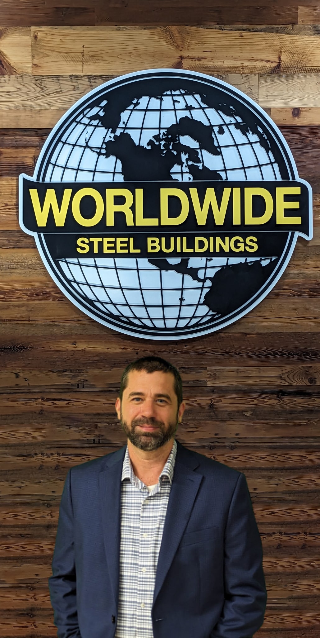 A person stands in front of a wooden wall with a sign displaying a globe and the text "WORLDWIDE STEEL BUILDINGS" in bold yellow and white letters. The person is wearing a checked shirt and a dark blazer.