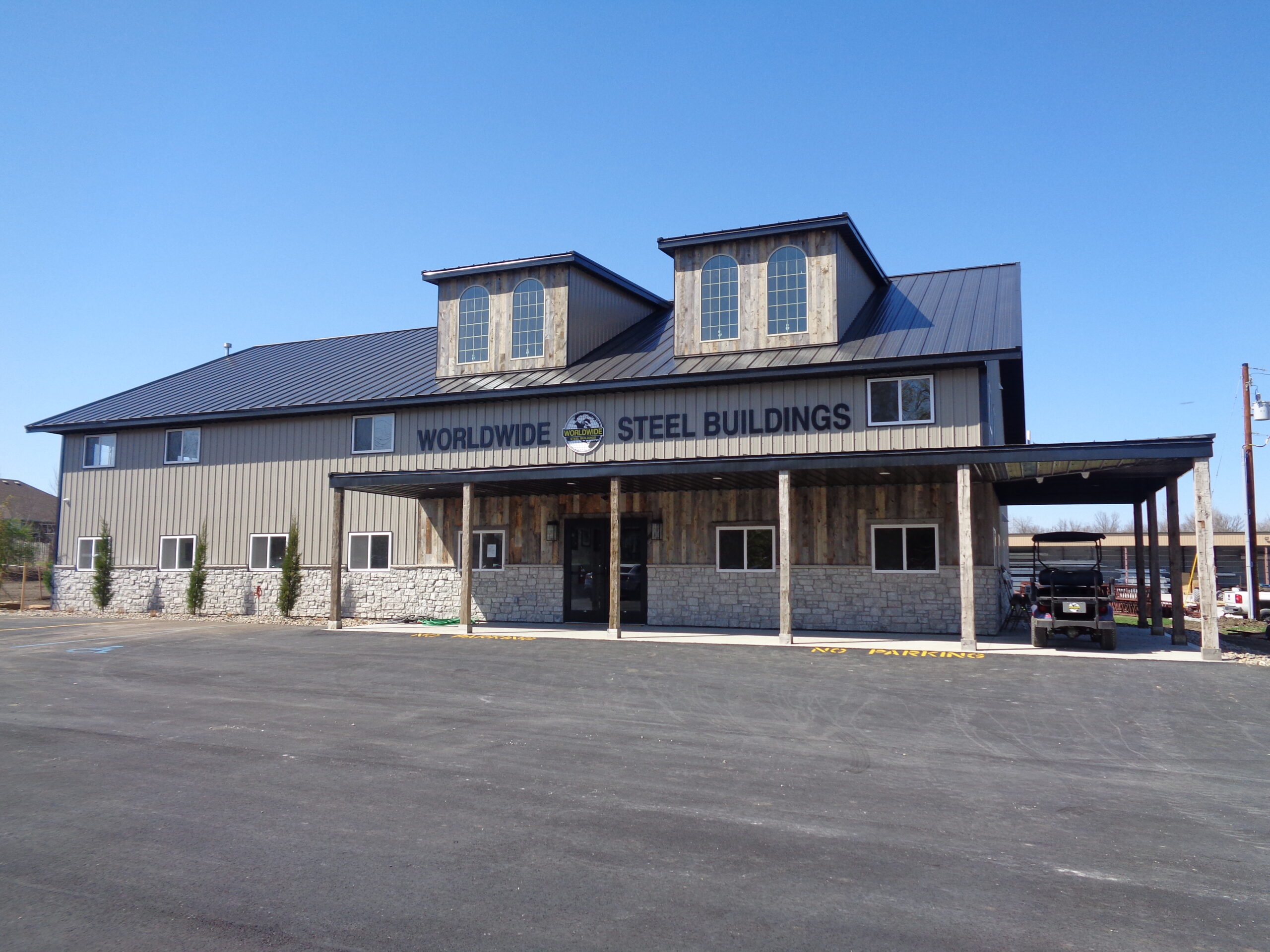 A two-story building with a metal roof and stone accents prominently displays the "Worldwide Steel Buildings" sign, representing a reputable steel building manufacturer. It features two dormer windows and a covered porch. In front, there is a paved parking lot with a small vehicle.