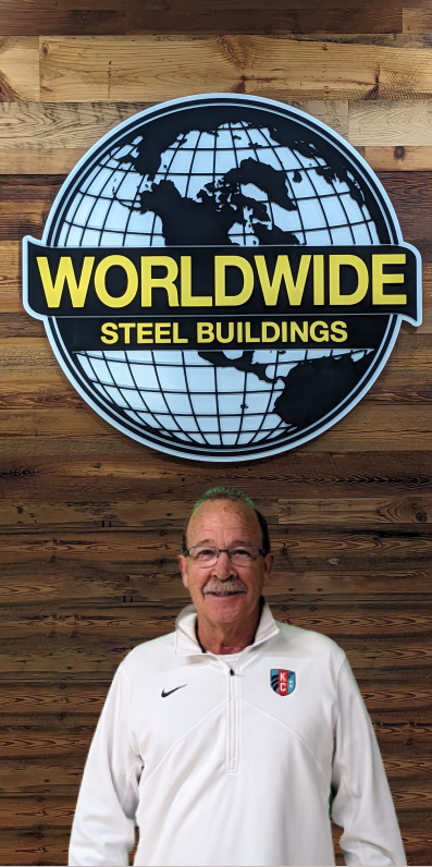 A man with a mustache stands smiling in front of a wooden wall. Above him is a large sign featuring a globe graphic with the text "WORLDWIDE STEEL BUILDINGS" in bold yellow and white letters.
