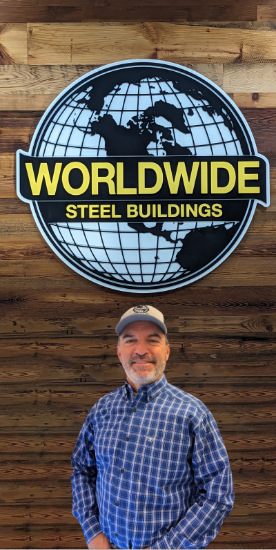 A person in a plaid shirt and cap sits smiling at a desk in an office, embodying the friendly spirit of the team. A computer monitor, papers, and a whiteboard with notes are visible in the background, illustrating their commitment to company goals.