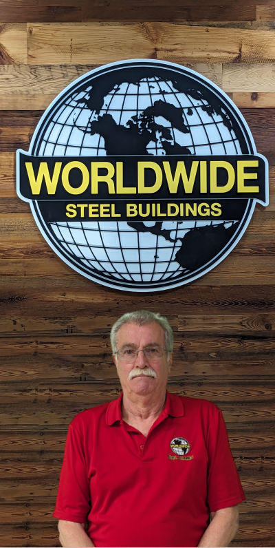 A man with glasses and a mustache is sitting at a desk cluttered with paperwork and a computer. He is wearing a beige polo shirt with a logo featuring the word "Builder." The background shows office supplies, a small framed photo, and colorful test buttons scattered around.