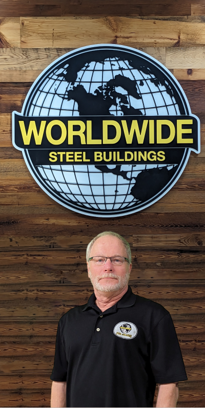 A man with glasses and light hair sits at an office desk, resembling a meticulous builder at work. He's wearing a black polo shirt with a logo that reads "State Aid Roads." Behind him are papers pinned to a board and a computer keyboard awaits his next button test.