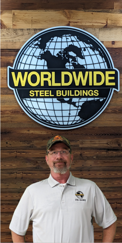 A man in a blue shirt sits at a desk with a computer displaying a spreadsheet. Office furniture and a wall calendar are visible in the background. With the poise of a seasoned builder, he looks toward the camera with a slight smile, ready to tackle any challenge at the push of a button.