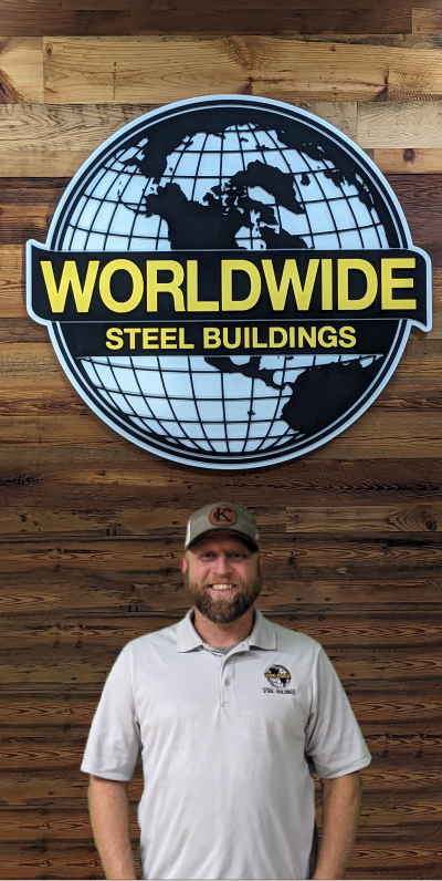 A person with a beard and a smile sits at an office desk wearing a red striped polo shirt. Behind them, a computer monitor and neatly stacked papers are visible, along with the builder's test plans. The workspace is organized and professional.