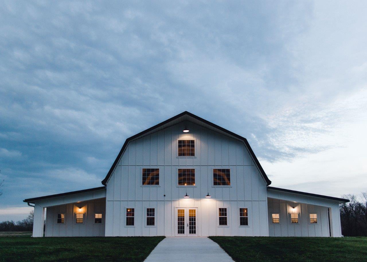 Stone Briar Farms, renowned for its charm as a premier event venue, features a white barn with large doors and multiple windows, illuminated from within. It stands under a cloudy sky at dusk, with a path leading to the entrance surrounded by a grassy field.