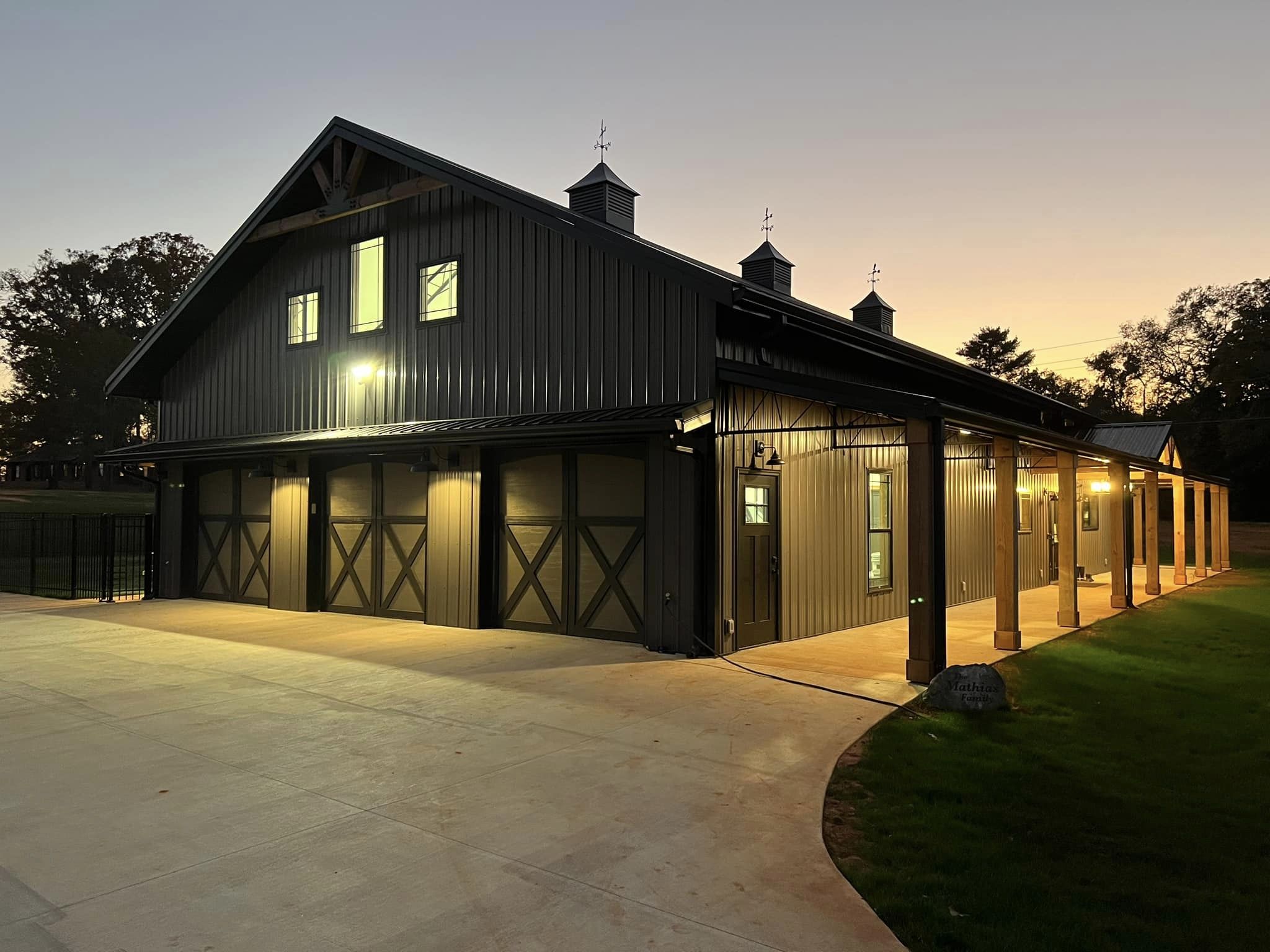 A large, modern barndominium is illuminated at dusk. It features multiple garage doors and a covered walkway. The clear sky and lighting cast a warm glow on the surrounding area, creating an evermore inviting atmosphere.