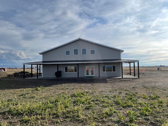 A large, two-story metal barndominium stands on a grassy Idaho field under a cloudy sky. The design features multiple windows and a covered porch that wraps around the front and sides.