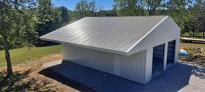 A large metal garage with a sloped roof stands on a gravel base in Missouri. It boasts two open garage doors and is surrounded by lush grass and trees under a clear blue sky.
