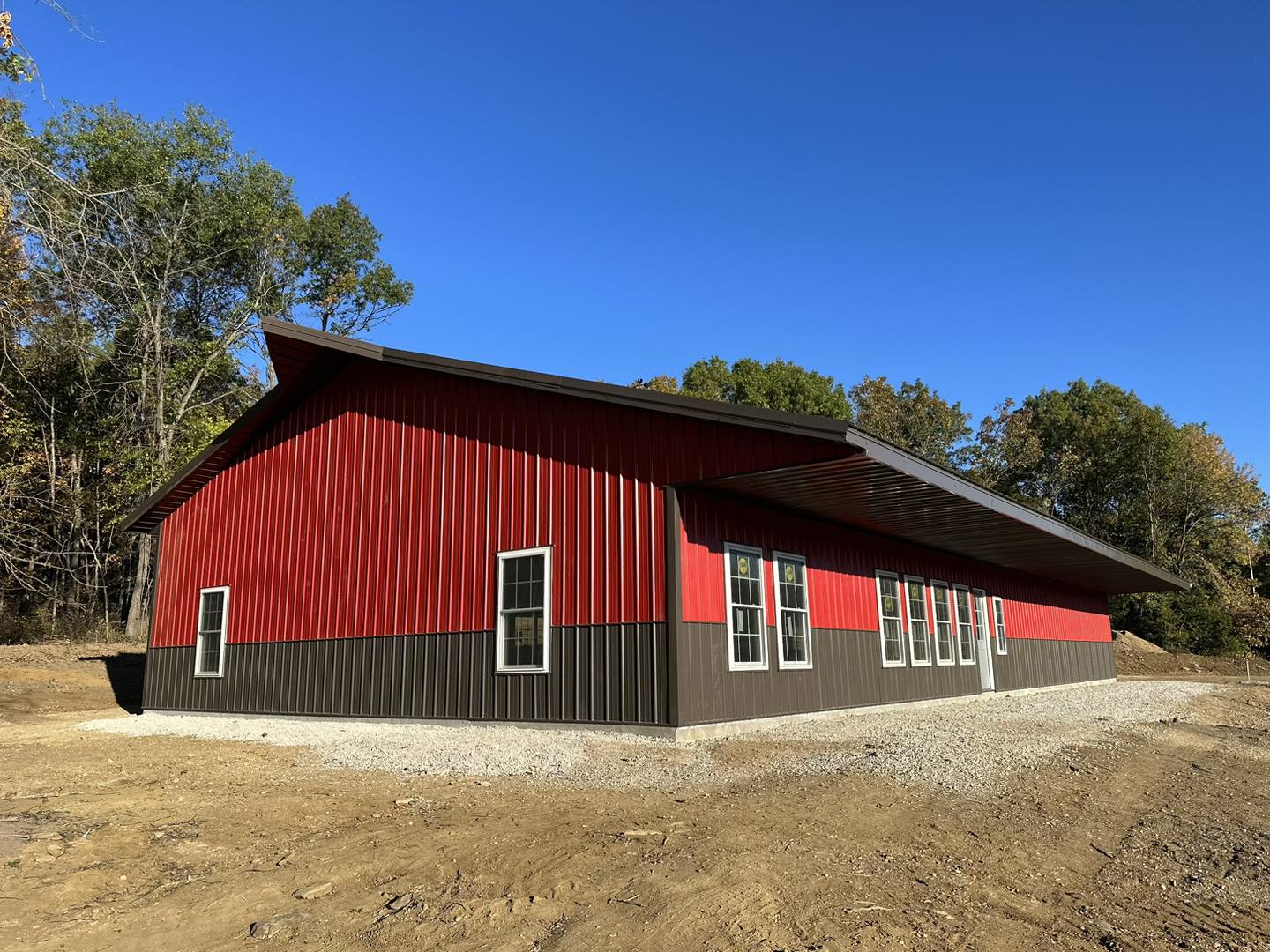 The red and brown Barndo with a slanted roof stands proudly on the cleared dirt area of DeLuca Farms. Surrounded by trees under a clear blue sky, it showcases several windows along its sides.