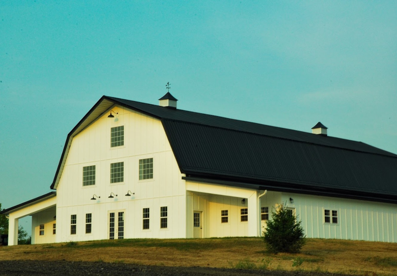 A large white barn with a black roof and cupolas sits on a slightly sloped grassy hill under a clear blue sky at Stone Briar Farms. The event venue features multiple windows and a weather vane atop, offering picturesque views of the surrounding landscape.