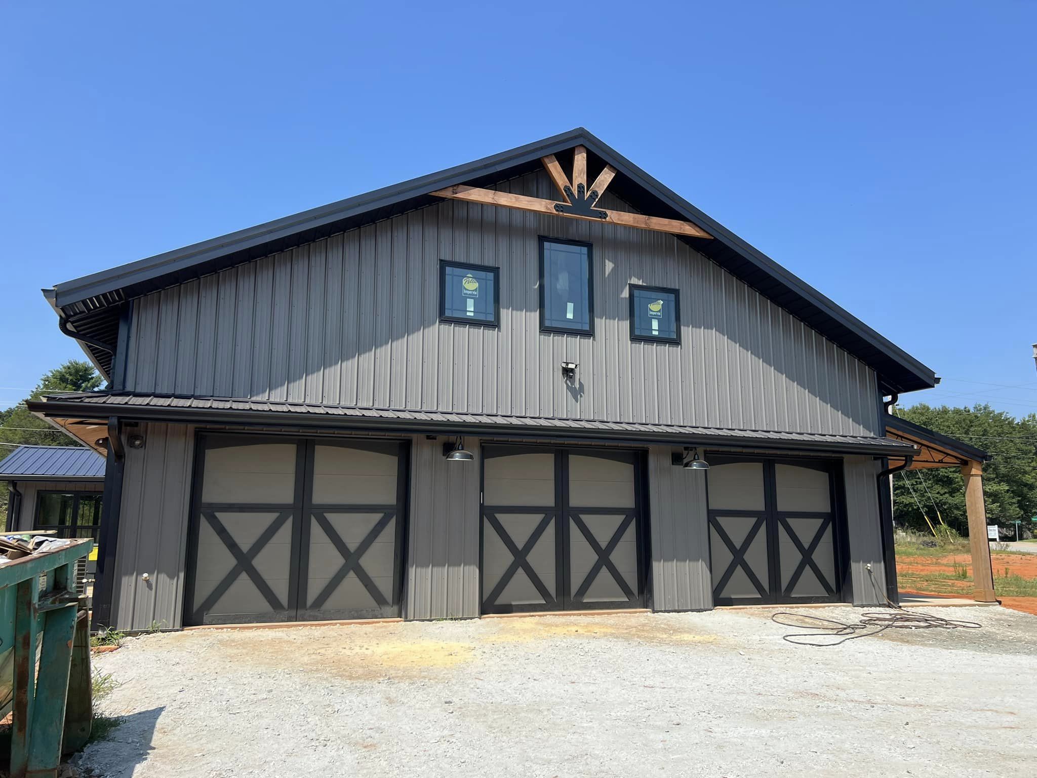 A large, gray barndominium with four garage doors showcases a decorative wood design at the peak. The dark roof contrasts with the gravel ground out front, while an evermore blue sky provides a stunning backdrop.