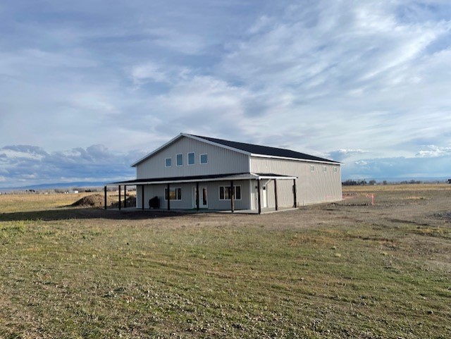 A large, metal-clad barndominium with a pitched roof stands on a flat, grassy area in Idaho. The sky is partly cloudy, casting soft shadows on the design. The landscape is open with distant hills and fields under a wide, blue sky.