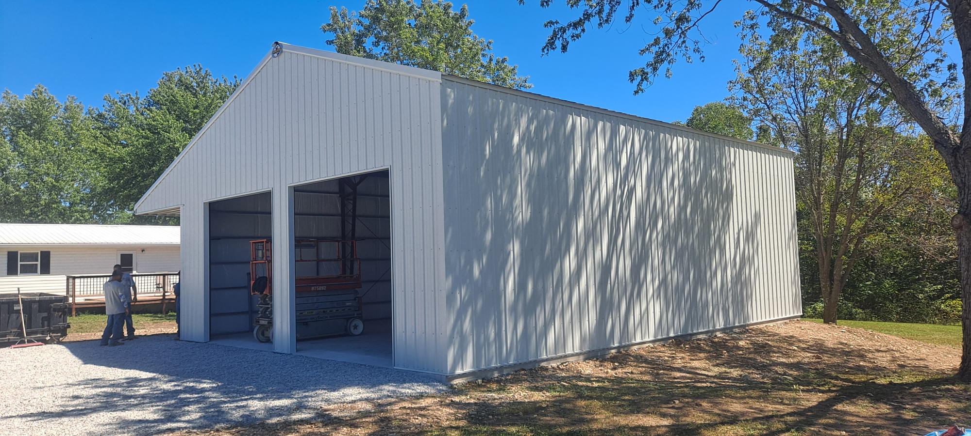 A large, white metal garage building with two open doors stands on a gravel area in Missouri, surrounded by trees and a nearby house. A person is visible near the entrance, and the scenery is under clear blue skies.