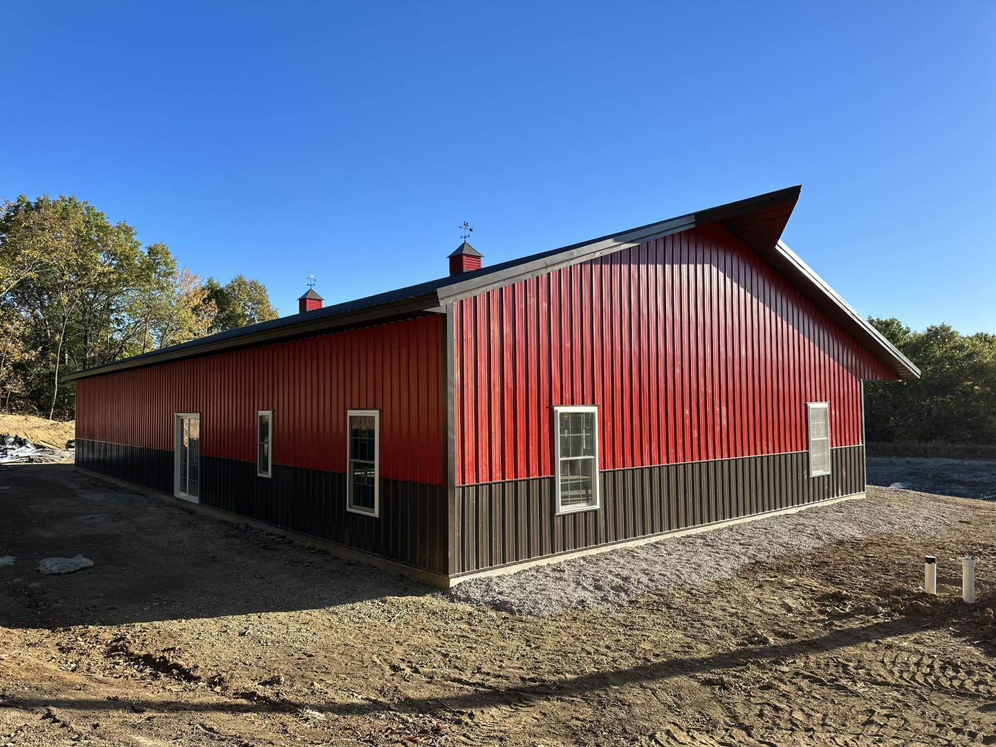 The red and brown barn, a classic barndo style, stands on a dirt patch at DeLuca Farms. It features a gabled roof adorned with two cupolas and several windows. In the background, trees frame the scene against a clear blue sky.