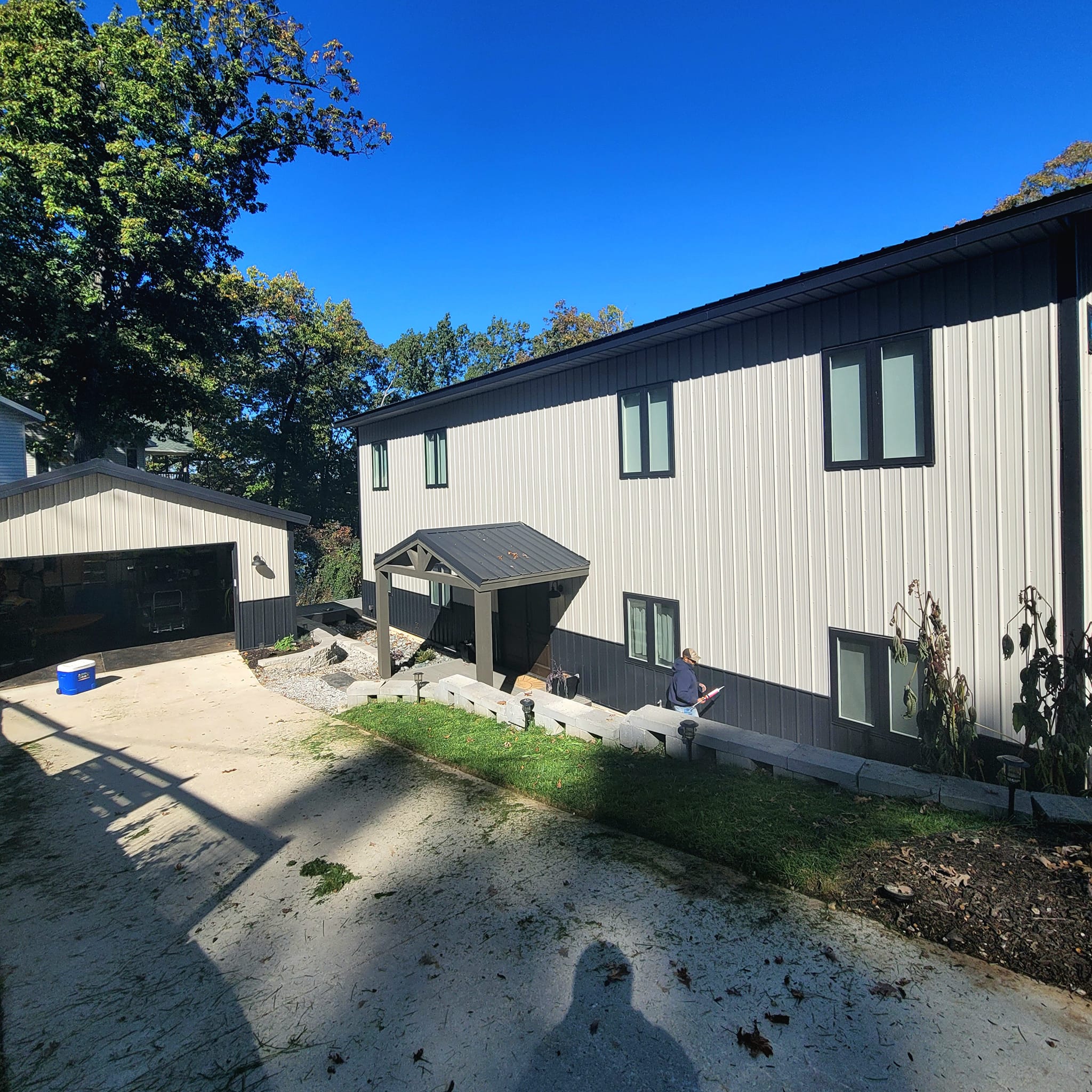 A modern, two-story Red Iron Lakehouse with light siding and contrasting dark trim sits under a clear blue sky, surrounded by trees casting shadows on the driveway. A small covered entrance and detached garage complete this picturesque setting near Table Rock.