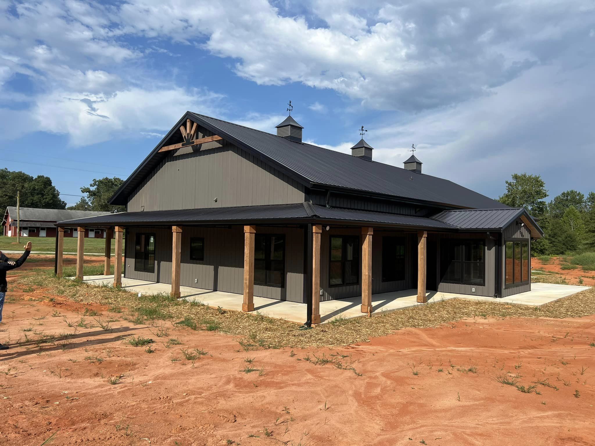 A contemporary barndominium with a black metal roof, wooden pillars, and expansive windows. It stands on evermore reddish-brown dirt under a partly cloudy sky, surrounded by sparse greenery. A person in the foreground gestures toward the building.