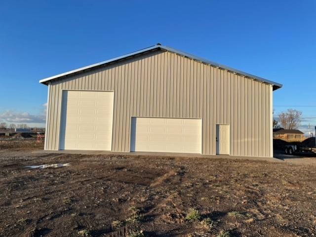 A large beige metal barndominium with three white doors—two garage doors and one regular door—stands majestically on an Idaho dirt lot under a clear blue sky.