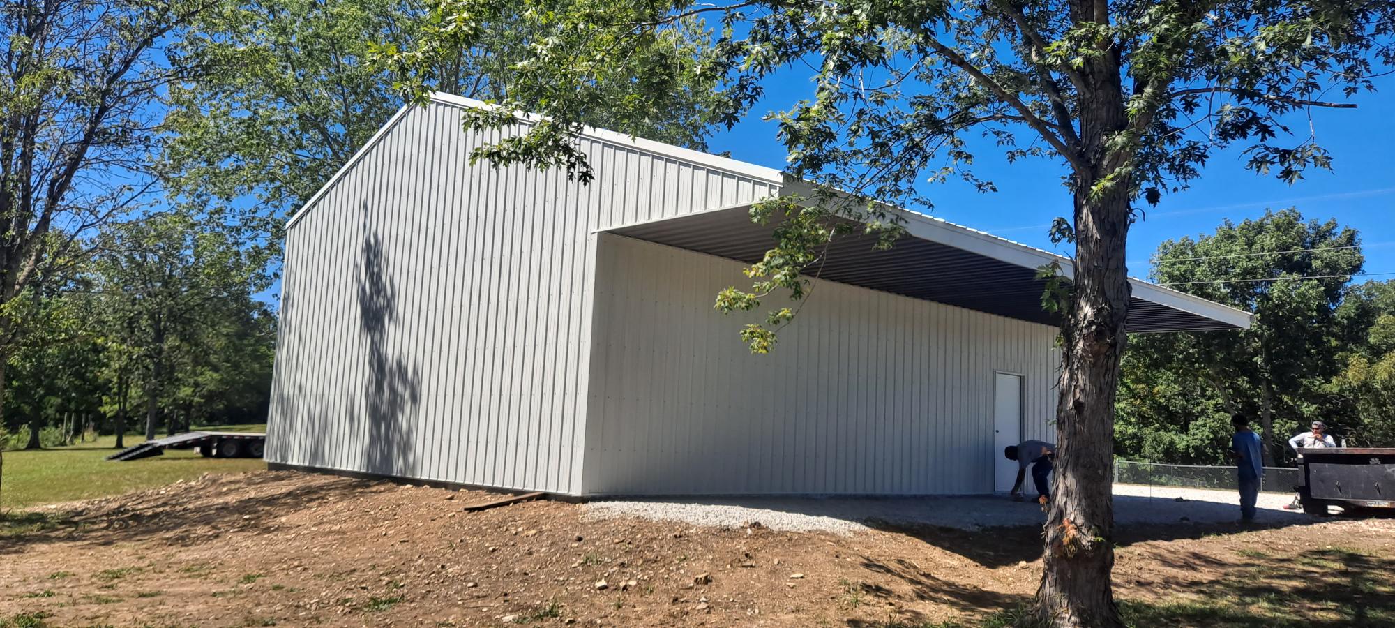 A large, light gray metal garage stands on a dirt and gravel surface, surrounded by trees under a clear Missouri sky. Several people are visible near the entrance, and the area is partially shaded by the building's overhanging roof.