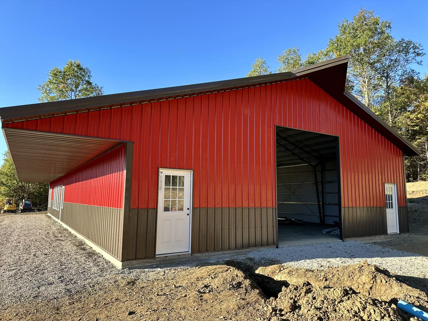 A large red and brown metal barndo with a sloped roof and two white doors stands on the gravel grounds of DeLuca Farms, surrounded by trees and a clear blue sky. One door is open, offering a glimpse into the interior.