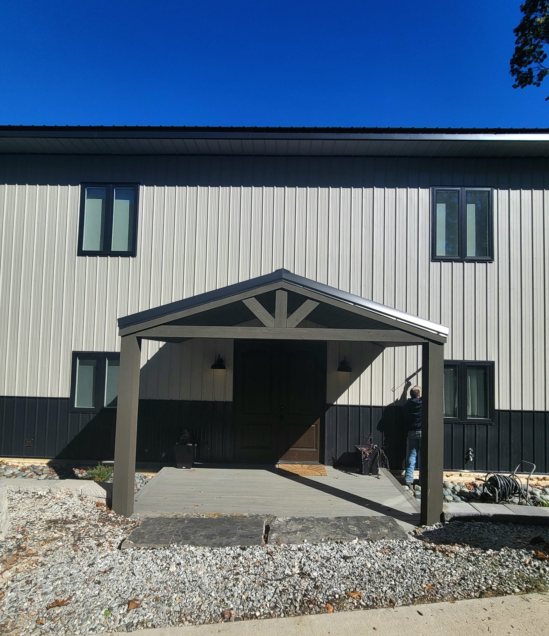 A modern two-story lakehouse with vertical paneling in beige and black colors. The entrance features a porch with a triangular roof, reminiscent of the scenic views at Table Rock. A person stands near the entrance under a clear blue sky, adding to the serene charm.