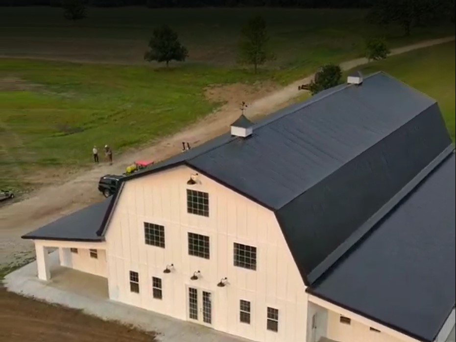 Aerial view of Stone Briar Farms reveals a large white barn with a dark roof, perfect as an event venue, nestled in a grassy area. A few people and vehicles are nearby. A dirt road winds through the landscape, with trees adorning the background.