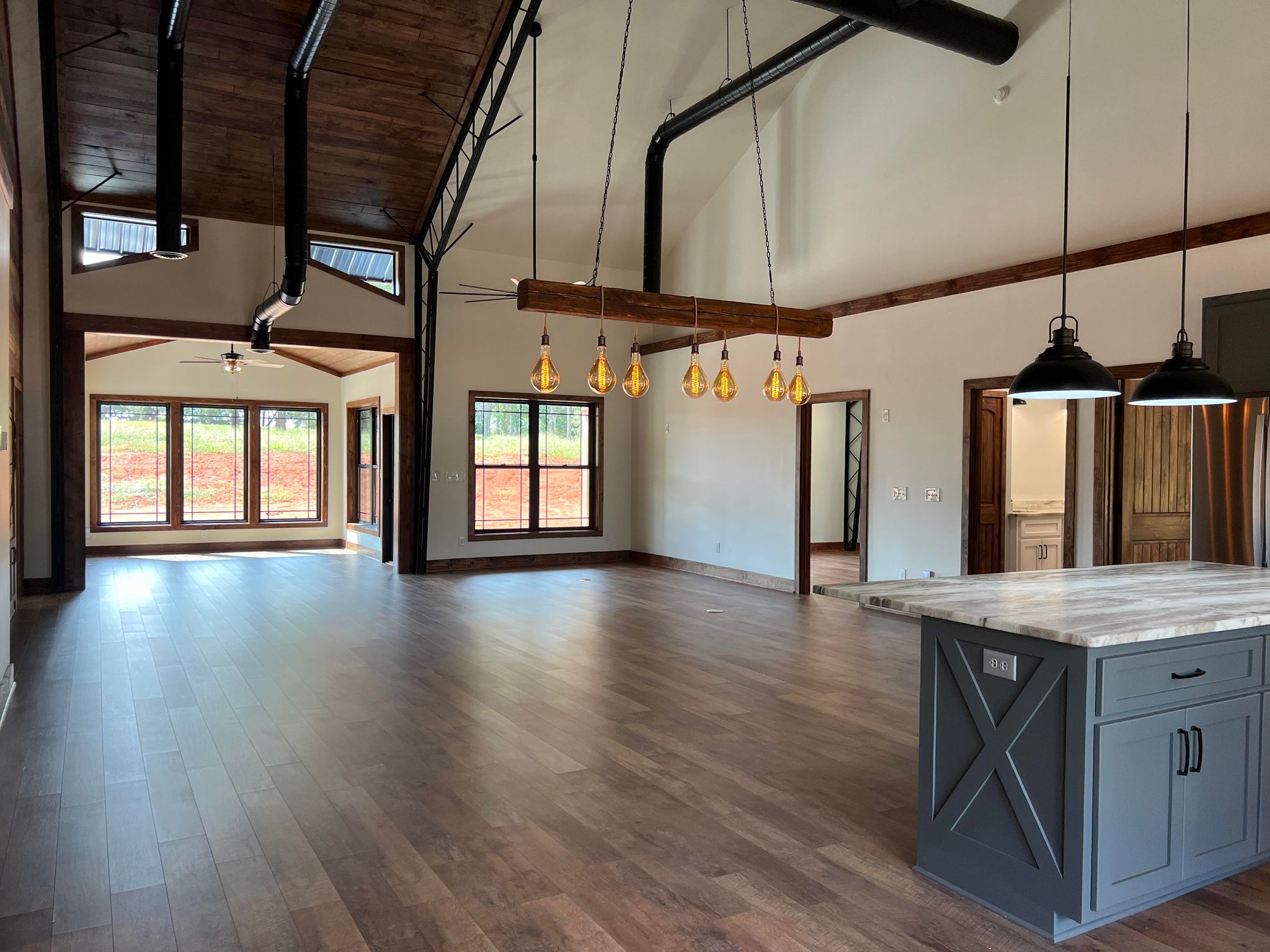 Spacious modern living area with hardwood floors, high wooden ceilings, and large windows exuding a barndominium charm. A kitchen island with a marble countertop and hanging pendant lights is visible in the foreground, perfectly capturing the essence of Evermore living.