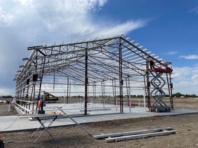 A partially constructed metal building on a concrete foundation, with visible beams and framework under a blue Idaho sky. A worker on a lift is on the right, with construction equipment and materials scattered around the site, suggesting the beginnings of an innovative barndominium design.