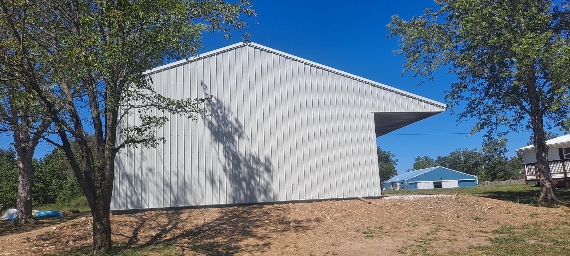 A large, modern light gray building with a slanted roof sits on a grassy area in Missouri. Trees frame the sides, and a smaller blue-roofed building, possibly a metal garage, is visible in the background under a clear blue sky.