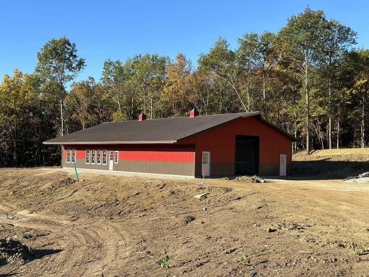 A red and gray barn with a black roof, typical of the Barndo style, sits on cleared ground at DeLuca Farms, framed by a forest with autumn foliage under a clear blue sky.