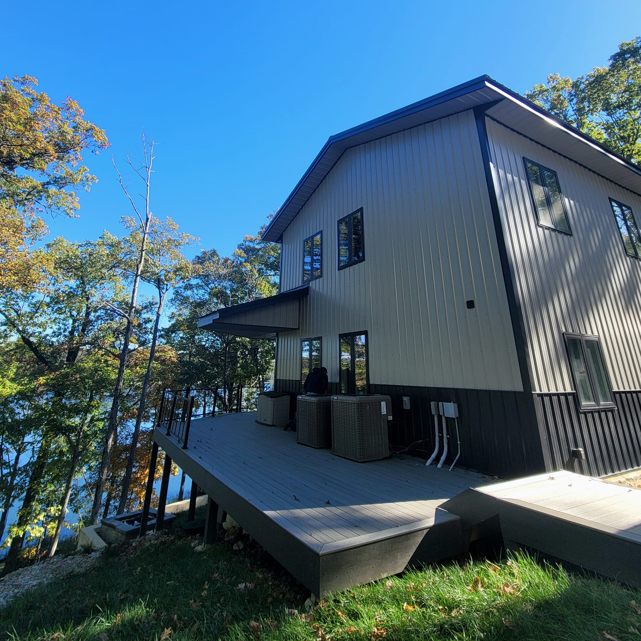 A two-story lakehouse with beige and black siding is situated on a raised wooden deck overlooking Table Rock Lake. Surrounded by trees with autumn foliage, the setting reflects a serene, natural environment under a clear blue sky.