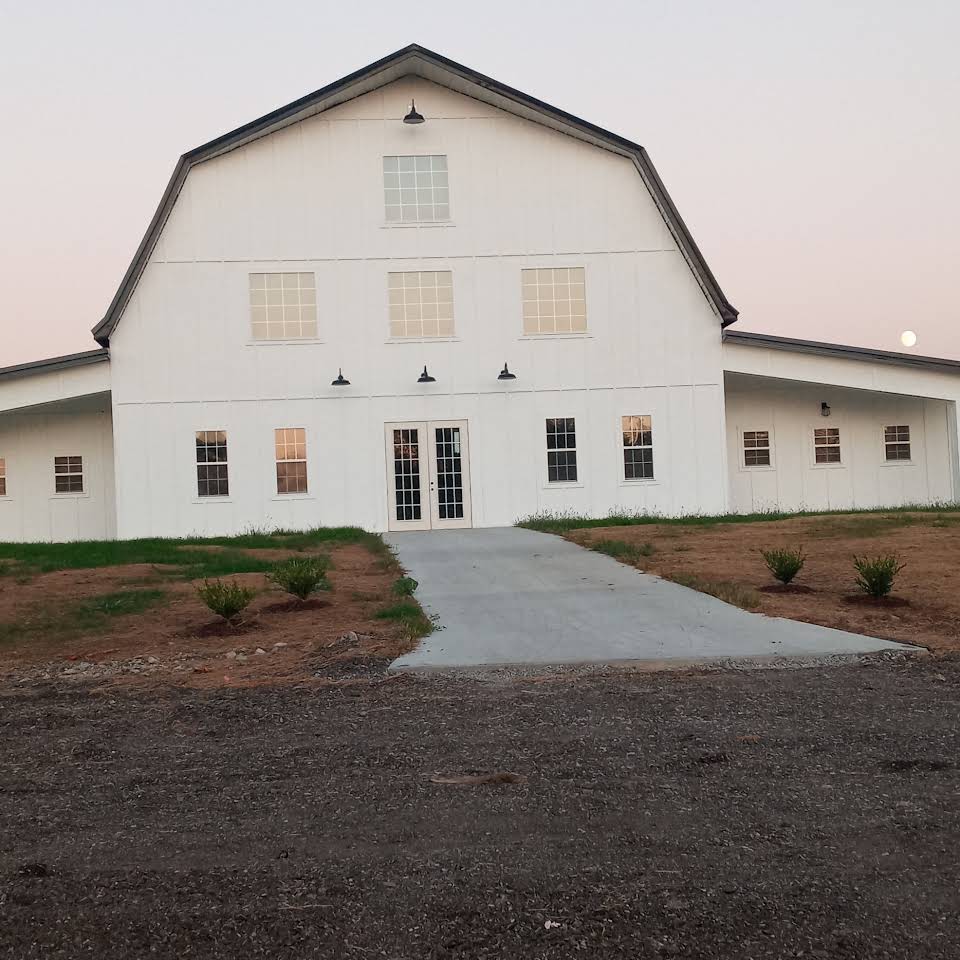 A large white barn at Stone Briar Farms boasts a steep roof and multiple windows, set against a dusky sky. A concrete path leads to the event venue's entrance, with small shrubs lining the walkway. Patches of grass are visible around the building.